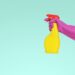 Aqua colored background with a hand wearing a magenta rubber glove, holding a yellow spray bottle