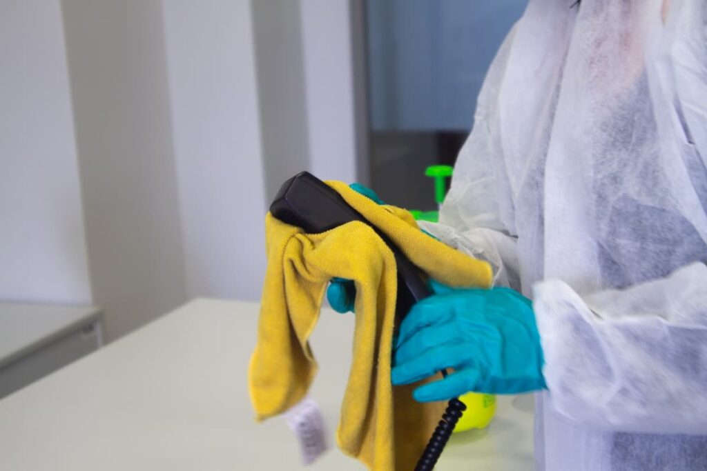 A person in white cleaning coveralls wearing blue gloves, using a yellow cloth to clean a black corded phone receiver