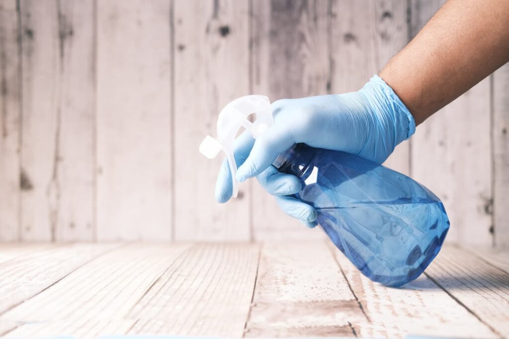Person wearing blue gloves spraying a spray bottle down on a wooden surface.